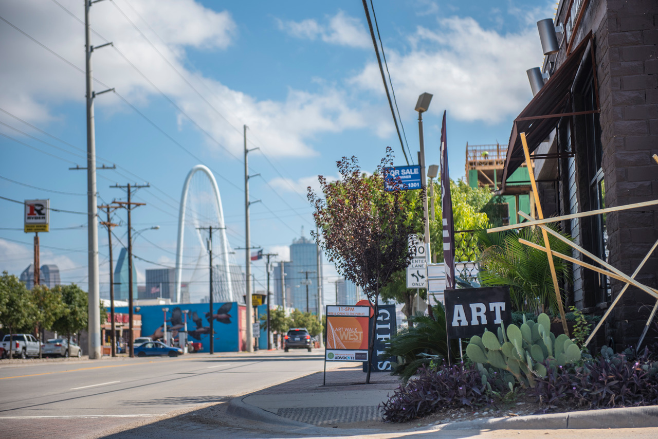 A street with trees, cacti and signs for various shops along the curb. In the background, there is a tiered arch bridge and the Dallas skyline.