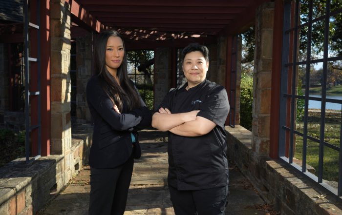 Two women, dressed in all black. Both have their arms crossed over their chest. The woman on the left is taller and has long hair, and the woman on the right has short hair. They are hosting a holiday fundraiser.