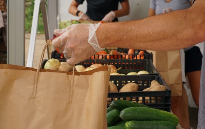 The hands of a volunteer at the dream center putting potatoes into a paper sack. In the background, a person sorts through vegetables.