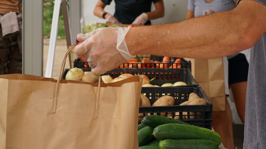 The hands of a volunteer at the dream center putting potatoes into a paper sack. In the background, a person sorts through vegetables.